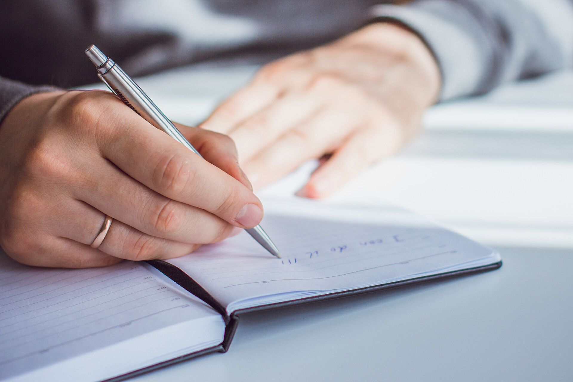 白色桌子上的人在写字一个男人的手像写着一句话："我能办到！""用圆珠笔写在记事本上"这个人坐在一张白色的桌子旁边，穿着一件灰色的夹克