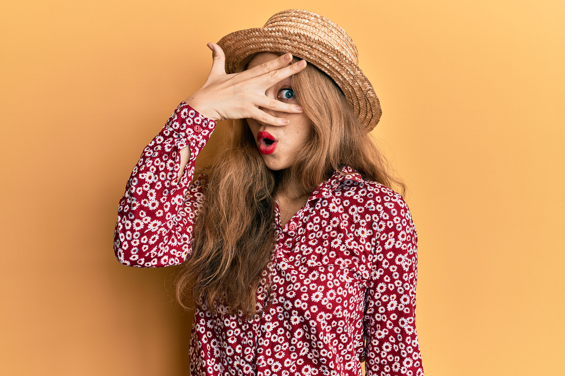 橙色背景前一个女人在拍照美丽的金发碧眼的女人戴着夏帽，满脸震惊，用手捂住眼睛，尴尬地看着手指. 