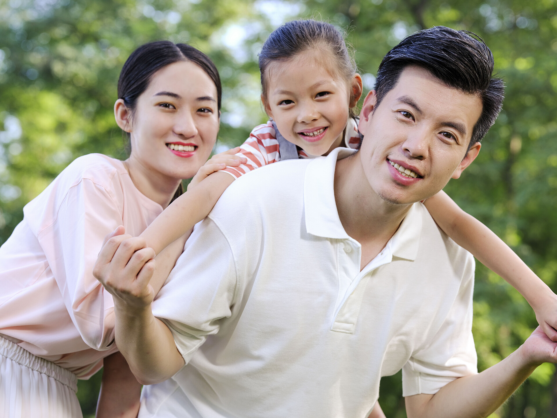 背着女儿的爸爸和扶着女儿的妈妈快乐的三口之家在公园里玩耍