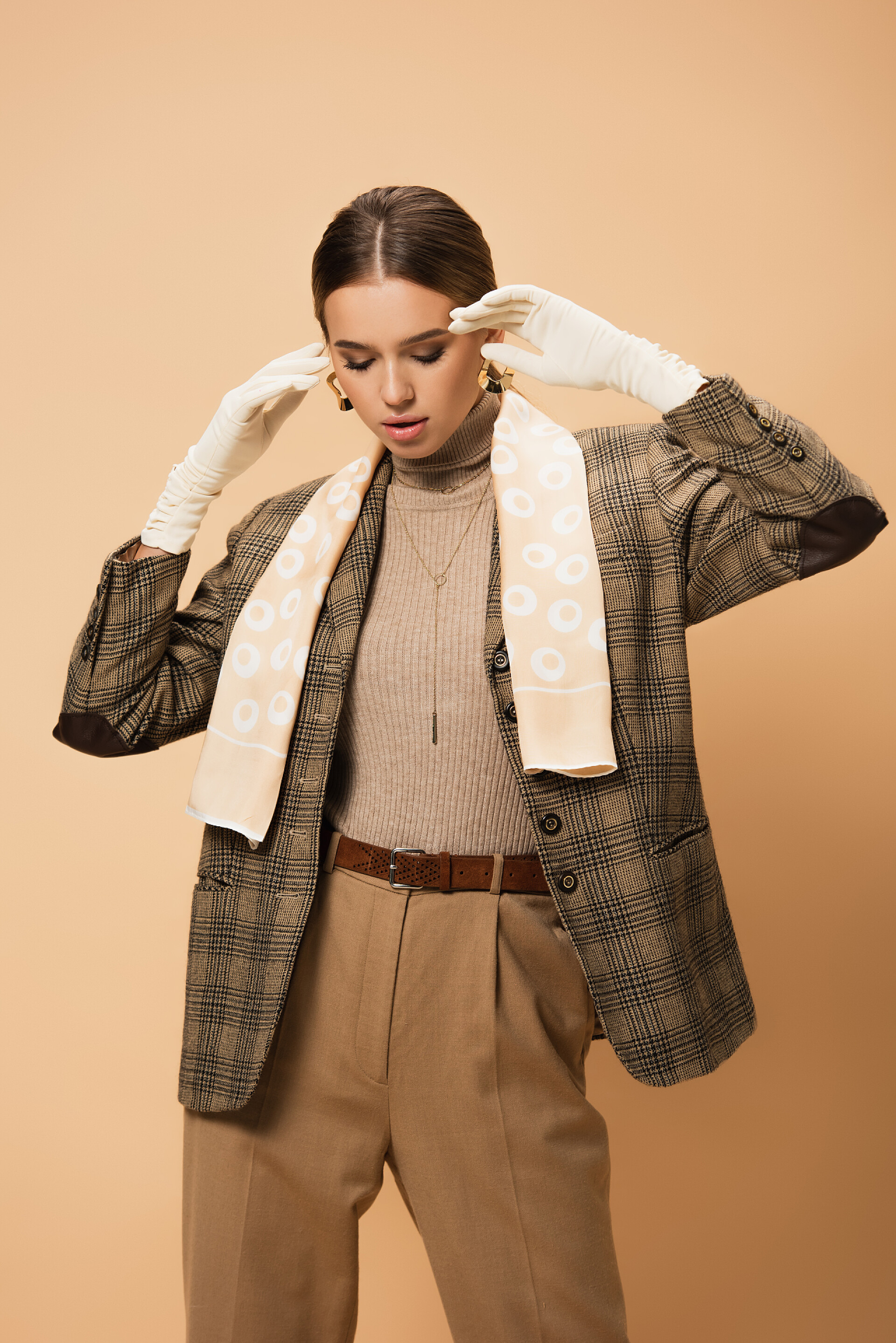 褐色背景下的一个时尚女人穿着格子呢夹克、戴着白手套、穿着米色背景衣服的优雅女人