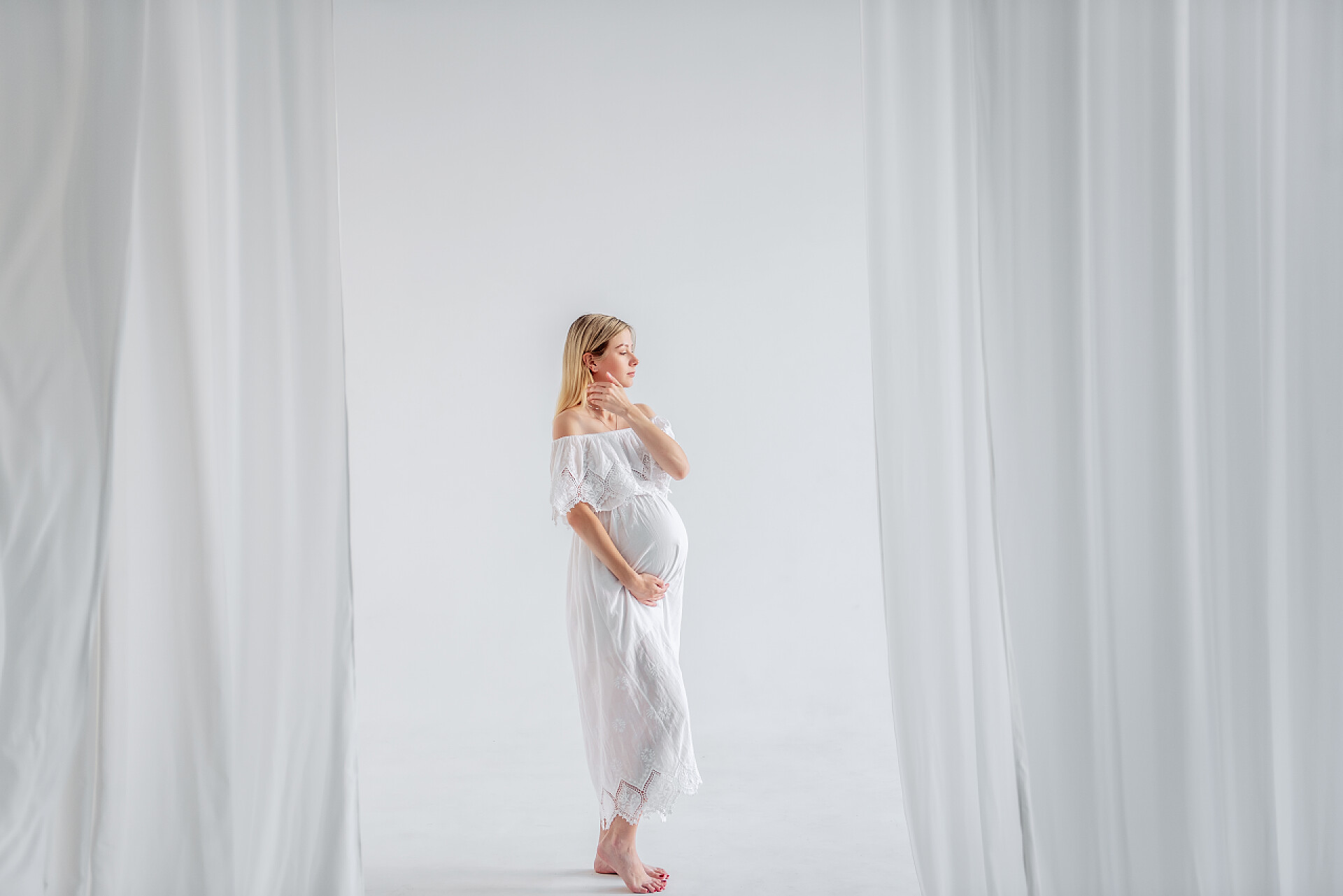 白色背景前一个怀孕的女人在拍照一个年轻的孕妇在期待着一个穿着白衣的孩子，抱着她的肚子。时尚女孩在一个简约的，孤立的内部削减空气的面料。容易、快乐的孕产、代孕.