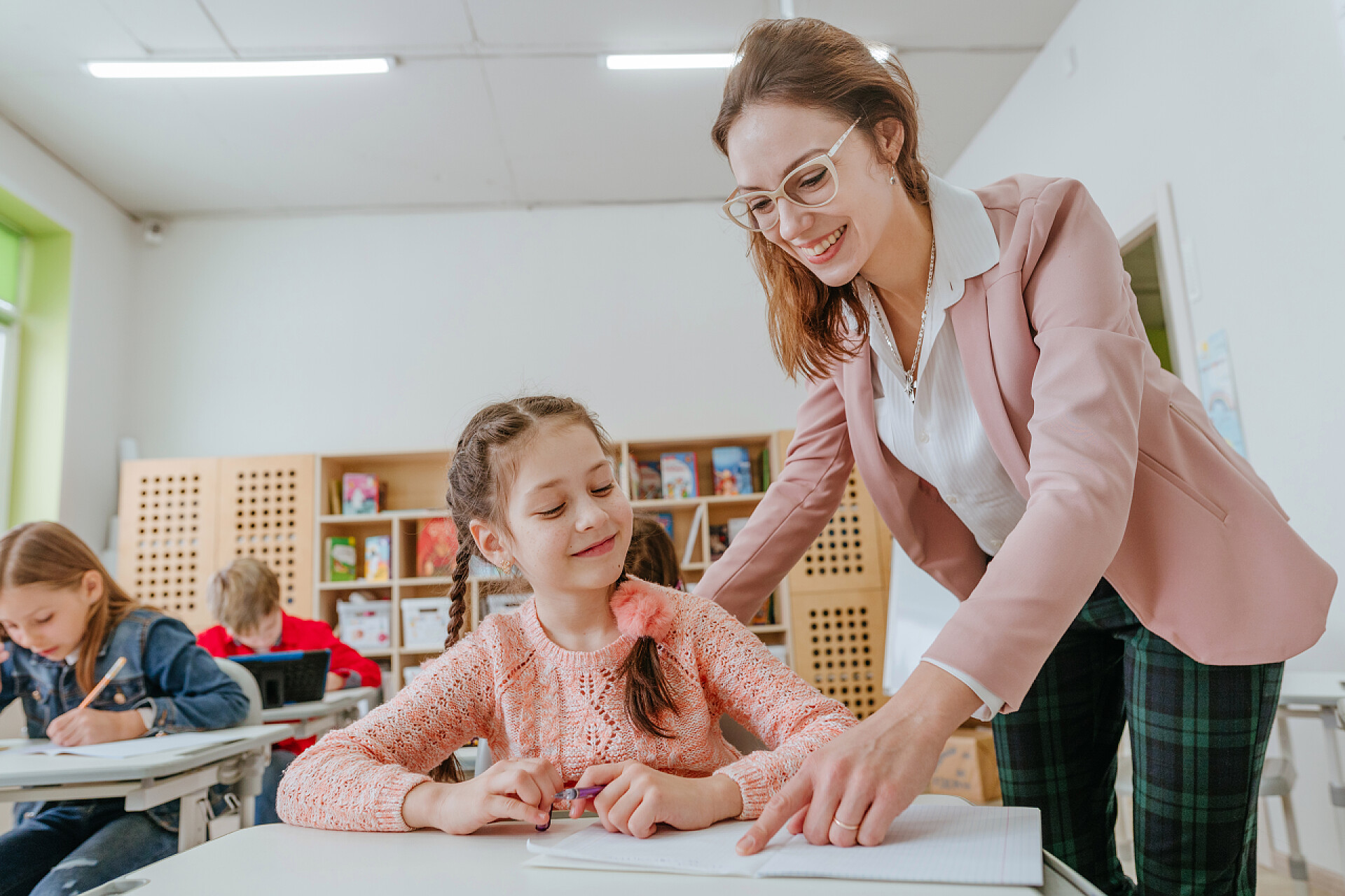 在班级里老师辅导小女孩学习女教师帮助女学生指着笔记本上的什么东西