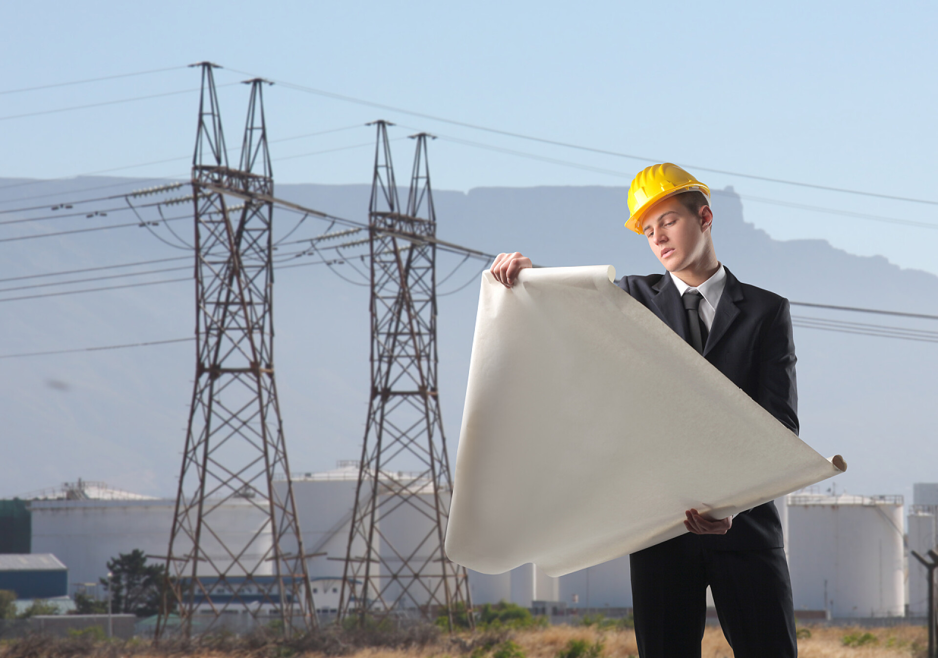在电网的草地上看图纸的工程师建筑工程年轻的工程师观测计划与工业网站的背景上
