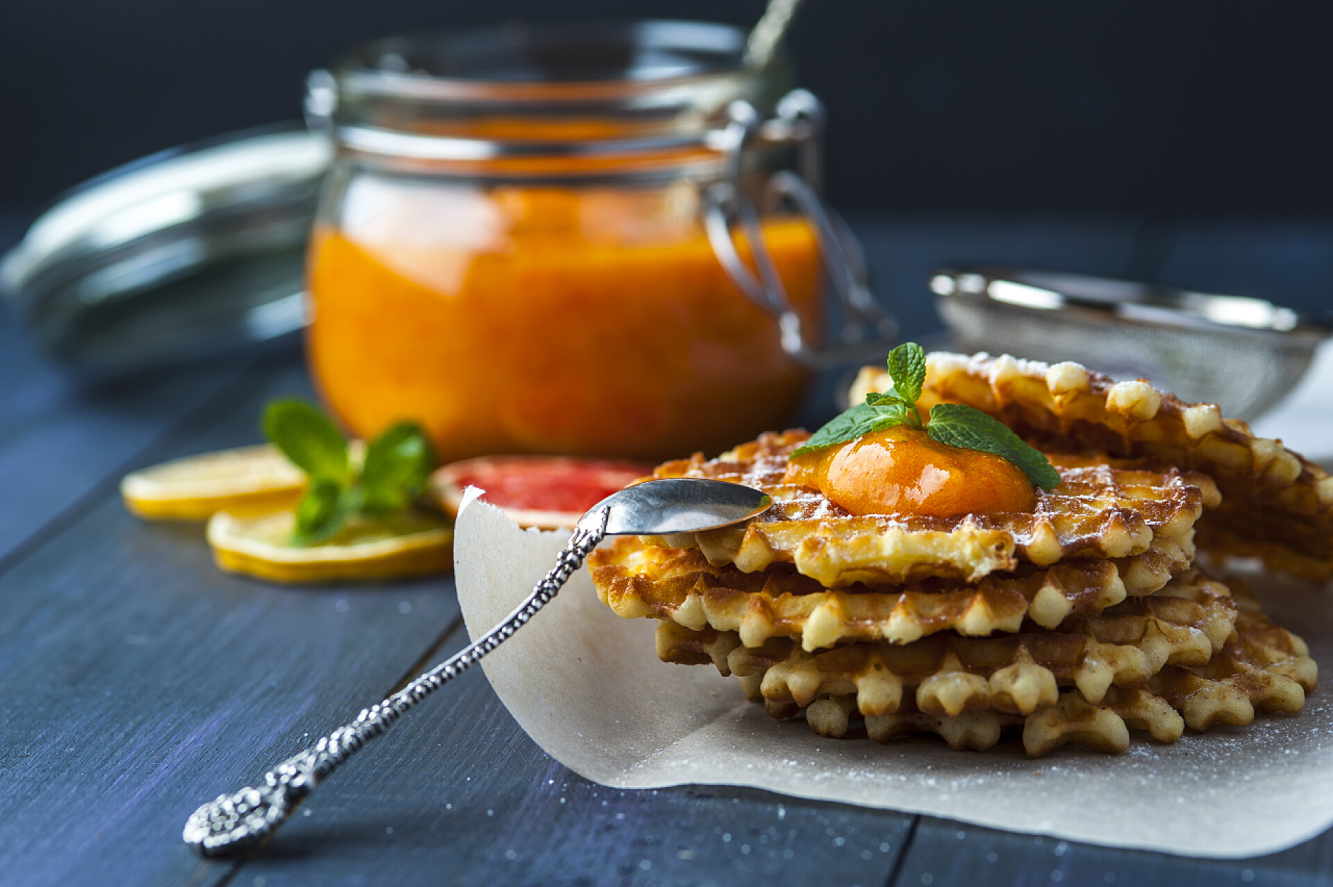 食品橘子酱曲奇饼干在烘焙纸上含银匙，一罐柿子果酱用勺子、柠檬和葡萄袖，飞碟在蓝色黑暗乡村背景小筛片晶圆。按横向的观点。专注于晶片.