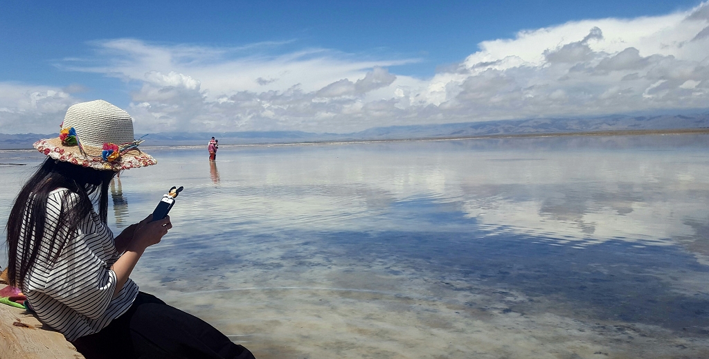 一位少女坐在茶卡盐湖岸边湖面上倒影着蓝天白云