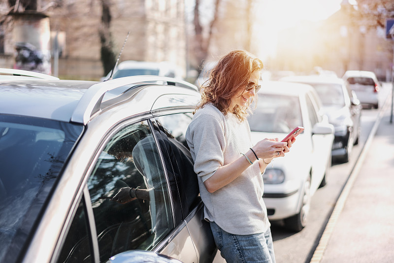 在城市的街道上，一名戴着墨镜的快乐的年轻女子站在她的灰色汽车旁，看着智能手机屏幕