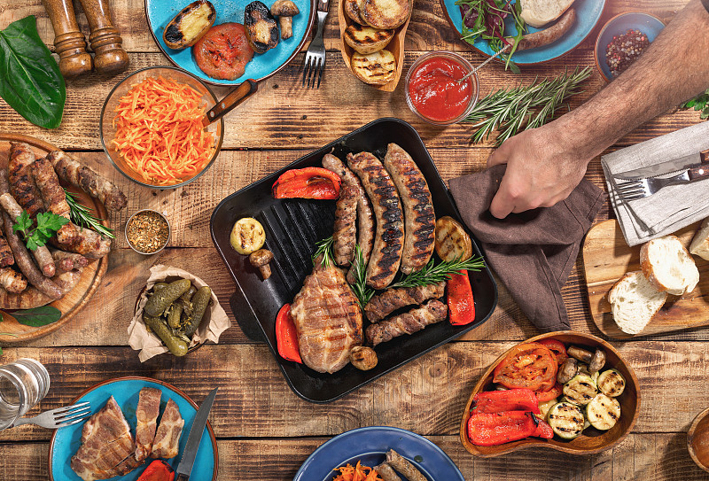 木桌上有不同的食物，烤牛排、香肠和烤蔬菜，俯视图