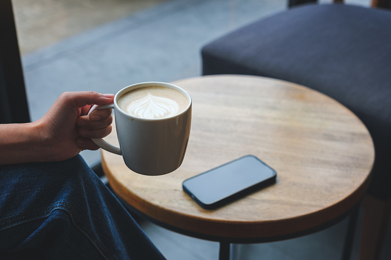 特写图像的一个女人喝热咖啡与手机在咖啡馆的桌子上