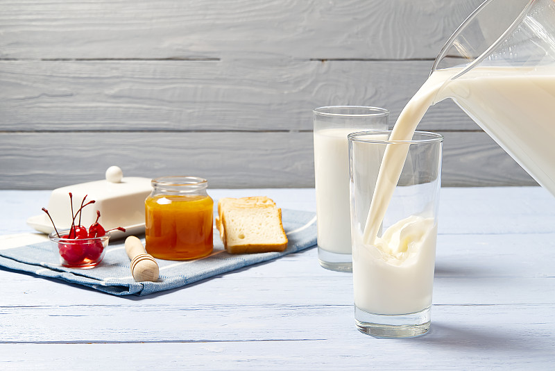 往玻璃杯里倒牛奶。一杯牛奶，蜂蜜和面包，浅蓝色的木头背景。乡村早餐食品概念。