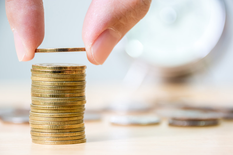 手放金币堆叠以模糊时钟为背景，金融投资省钱理念