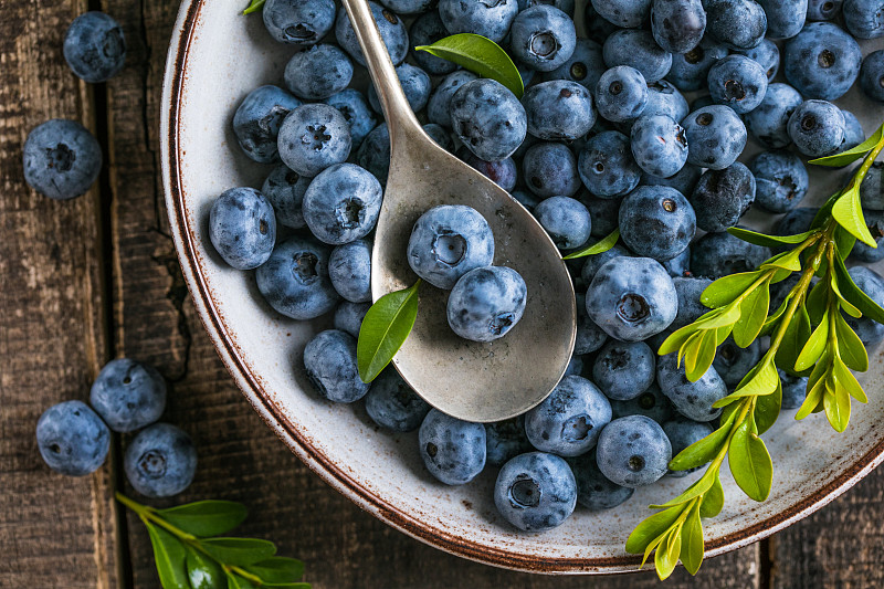 碗里新鲜采摘的蓝莓。多汁的新鲜浆果和绿叶放在质朴的餐桌上。健康食品和营养观念