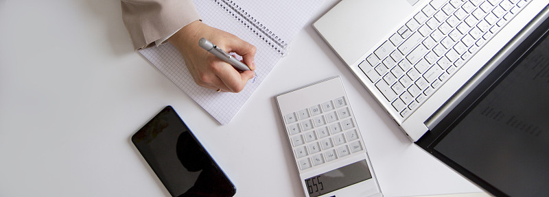 女商人用计算器和笔记本电脑计算预算，并在桌面上的笔记本上做笔记。