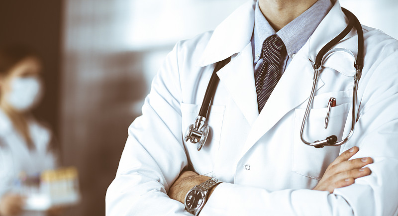 不明身份的男医生戴着防护口罩，双臂交叉站在诊所里。冠状病毒大流行时期的医学理念。Covid 2019