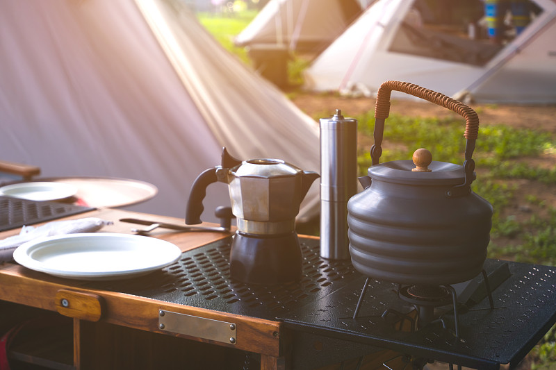 露营桌上的老式户外黑壶、咖啡研磨机和户外厨房设备，清晨阳光照在露营区帐篷表面