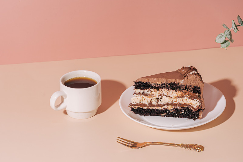 一块蛋糕放在白色盘子上，旁边放着叉子，一杯咖啡放在粉红色的桌子上