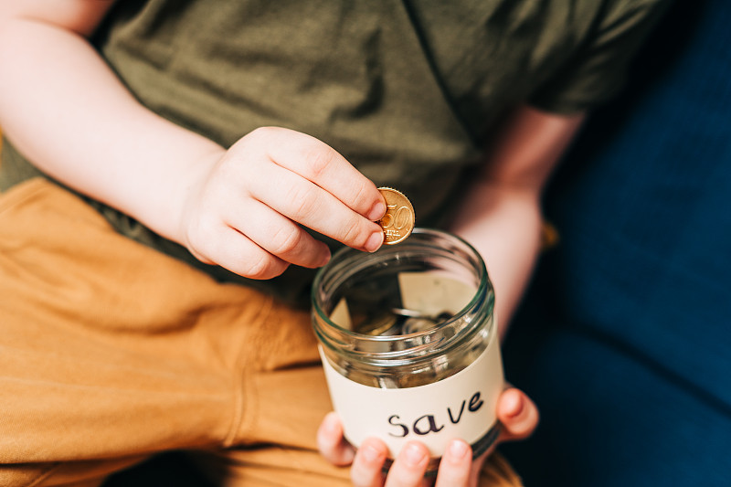 近距离的小男孩的手抓和把堆栈硬币在玻璃罐与保存标签。捐赠、省钱、慈善、家庭理财计划理念