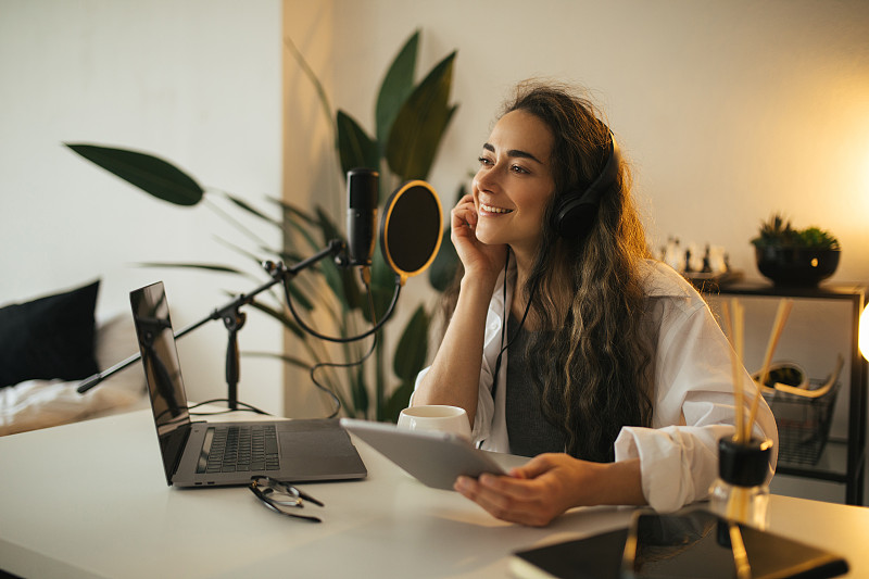 一名留着黑色长发的年轻博主在录音棚里用笔记本电脑、耳机和专业麦克风录制在线播客。