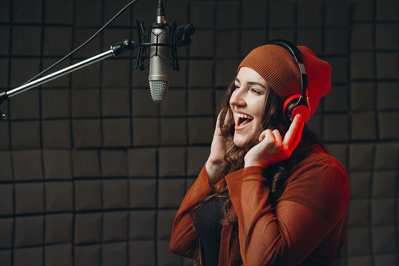 一名戴着耳机的年轻女子在录音棚里对着麦克风深情歌唱。明星，才艺，准备表演，录音棚录音。专业录音隔音室