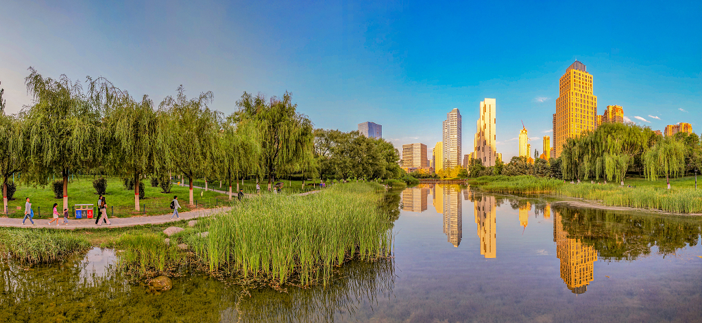 傍晚霞光照亮了高楼大厦并倒影在河水中河岸边小路上市民在散步