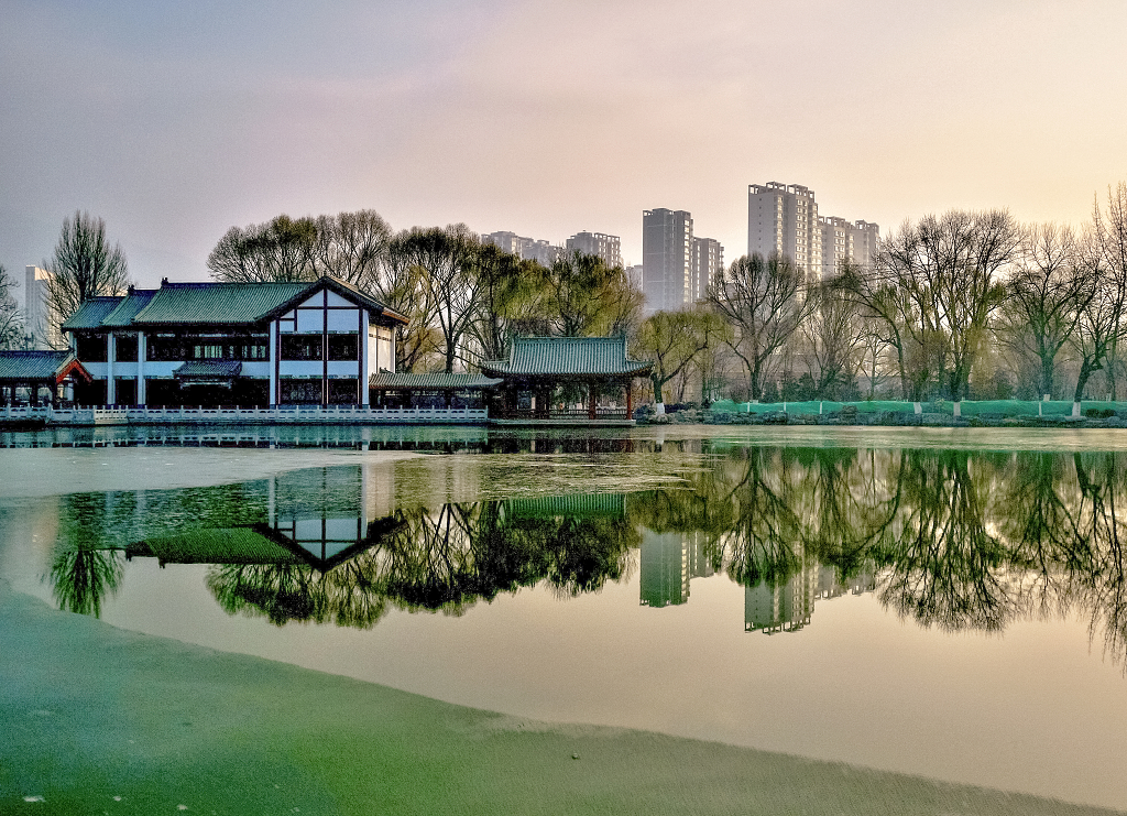 初冬的日出时分公园的湖岸边的房屋树木倒影在水面上