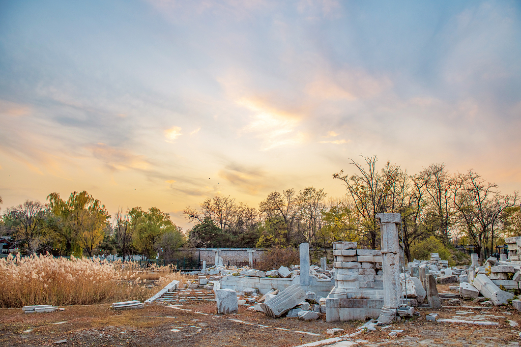 晚霞时分天空五颜六色地面上被破坏的圆明园残骸和树木