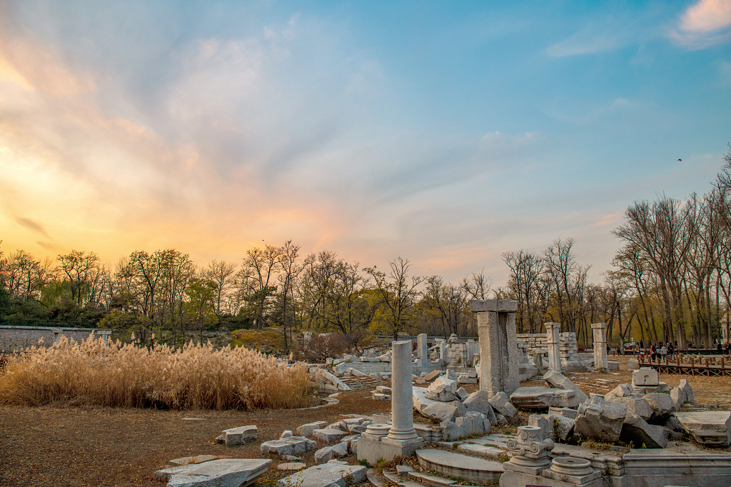 晚霞时分天空五颜六色地面上被破坏的圆明园残骸和树木