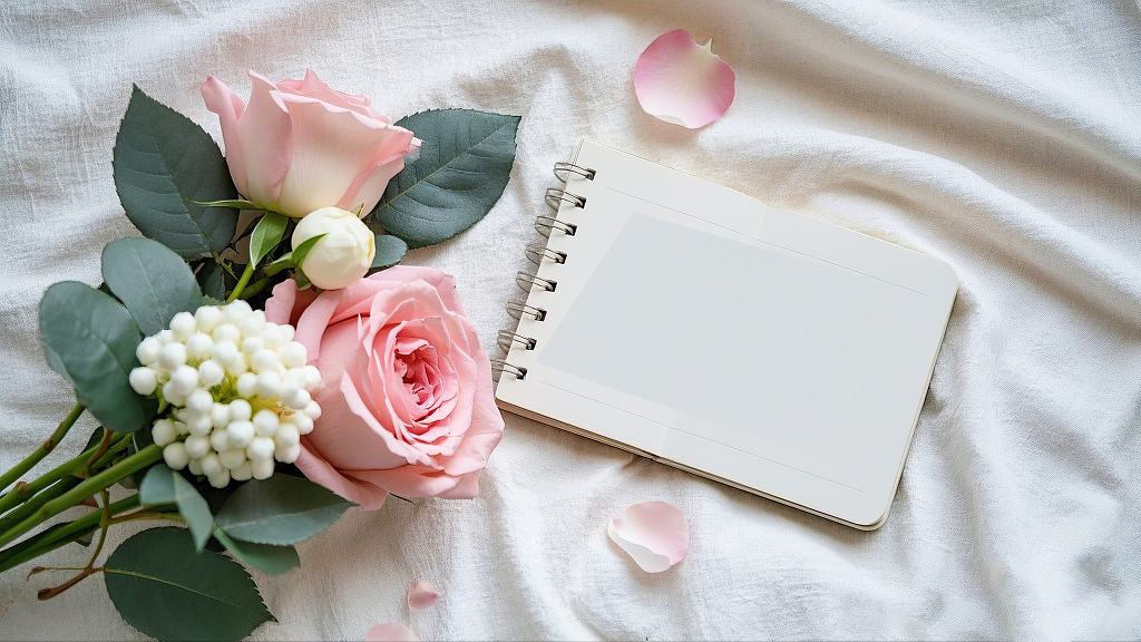 浅色布上摆放着粉色玫瑰白色小花和空白笔记本	AI