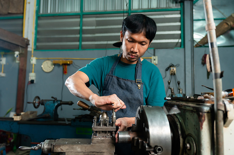 特写的亚洲年轻人的脸，严肃的表情控制，并在一个车床，在他的小工厂，这是一个家族企业。