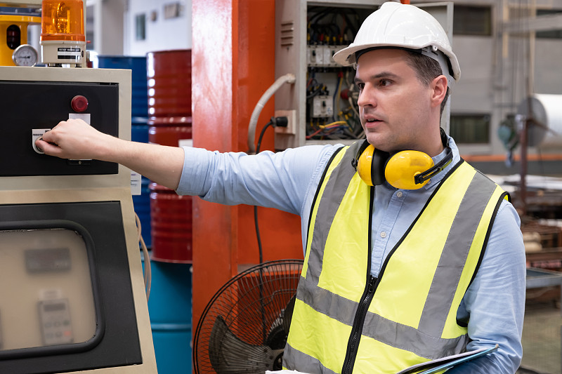 择优关注白人男性机械工程师，穿戴安全装备。同时负责厂区内机器的维护和安全控制。带有模糊背景的重型机械。