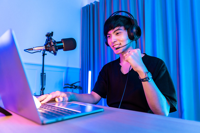 用智能手机玩游戏。年轻的亚洲帅哥坐在椅子上，手里拿着手机。霓虹灯室戴着耳机在线手机游戏退出流媒体。在线电竞流媒体游戏，家庭隔离。活动。