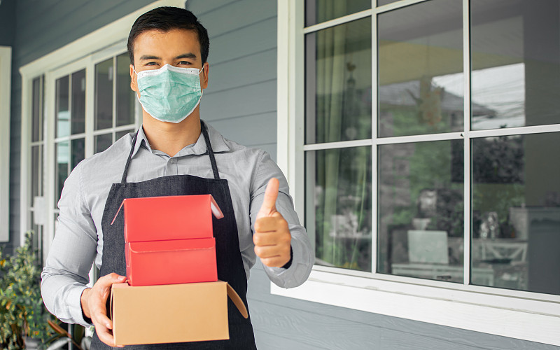 白人男性咖啡店和面包店老板，企业家戴着口罩保护病毒，拿着送货的盒子，站在商店外面。新常态，社会距离概念。