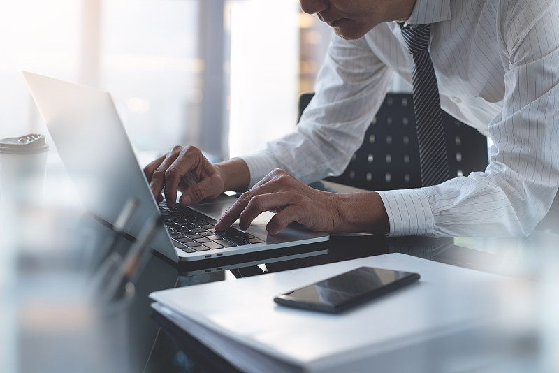 亚洲商人在现代办公室的玻璃桌子上用笔记本电脑工作