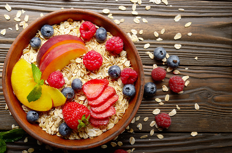 在厨房的木桌上的粘土盘子里放着桃子、草莓、杏仁和黑莓等健康的水果什锦麦片