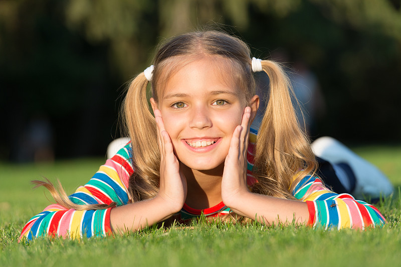 快乐微笑的学生。玩得开心。女孩可爱的孩子躺在绿色的草地上。健康的情绪，快乐的孩子在户外放松。什么让孩子快乐。女孩梳马尾辫，享受放松。快乐的女生