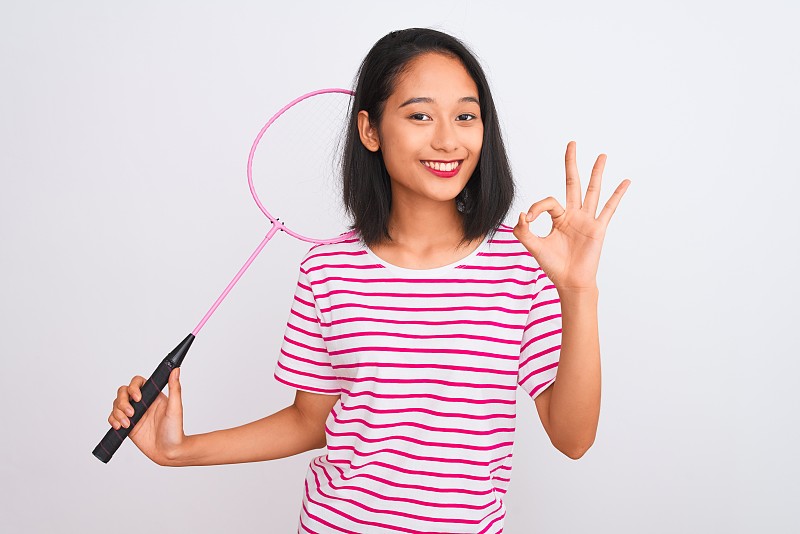 年轻美丽的中国女子拿着羽毛球拍在孤立的白色背景上用手指做ok手势，优秀的象征