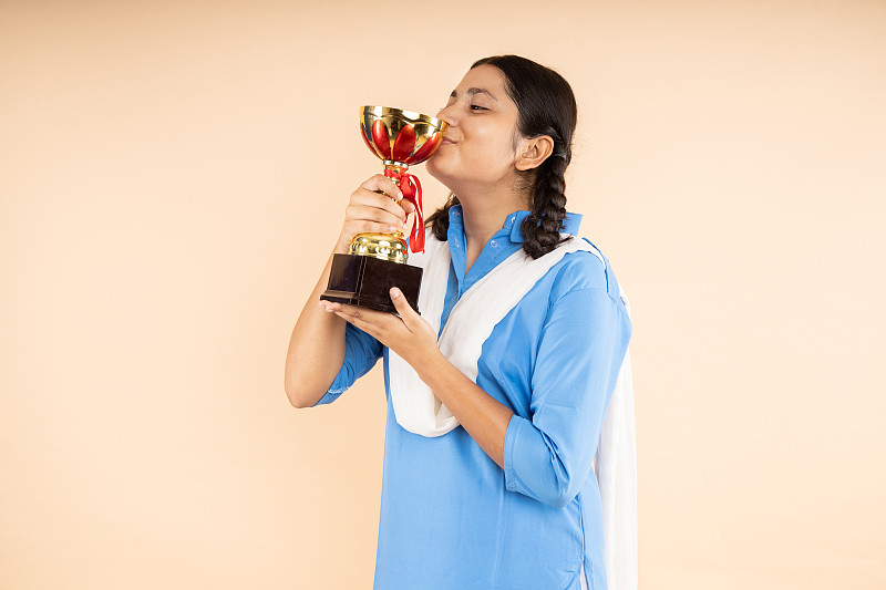 快乐的农村印度学生女学生穿着蓝色政府校服举行亲吻胜利奖杯在米色背景，获胜者，工作室拍摄，特写，教育概念。