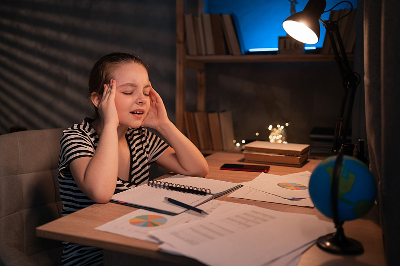 哈欠连天的十几岁女孩做作业在桌子在晚上，过度工作，筋疲力尽的作业女学生坐在一张桌子。厌倦了学校。