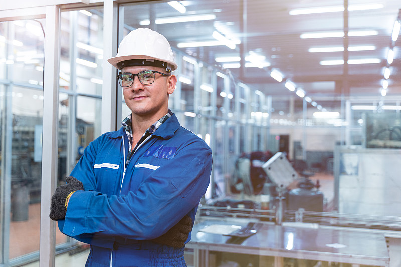 一幅手工工人的肖像正自信地站在高科技洁净工业工厂的玻璃墙前，穿着蓝色工作服，戴着安全帽。智慧产业工作者运作理念。