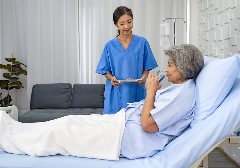 一位亚洲白发老人从护士手中接过药和一杯水。一位老妇人躺在医院的病床上接受治疗。