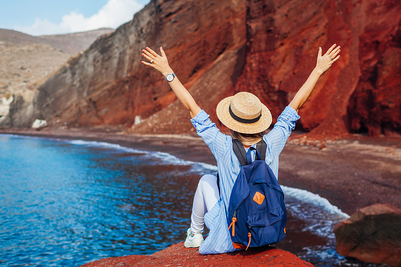 在希腊圣托里尼岛的红海滩上，女游客在岩石上放松地举起手臂。旅行和度假