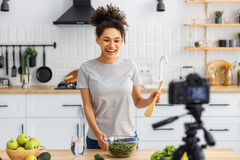 健康的生活方式。美丽的女人用相机为他的追随者们录制烹饪健康食品、或晚餐的视频。非裔美国年轻女性在家中厨房准备沙拉
