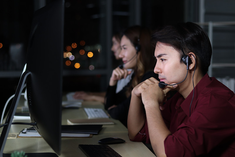 快乐微笑的亚洲人晚上在呼叫中心服务台顾问与队友一起工作，客户服务主管深夜带着麦克风耳机使用电脑支持办公室。