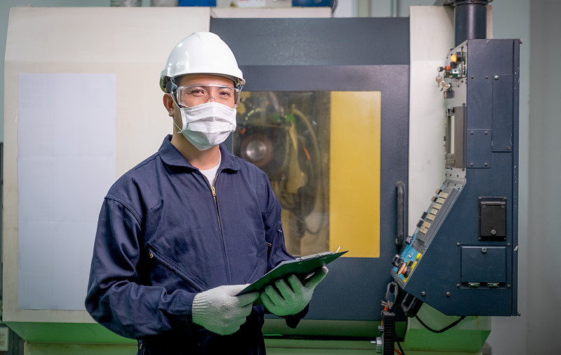 工厂工人戴着口罩拿着文件，也拿着文件文件，站在操作机器前