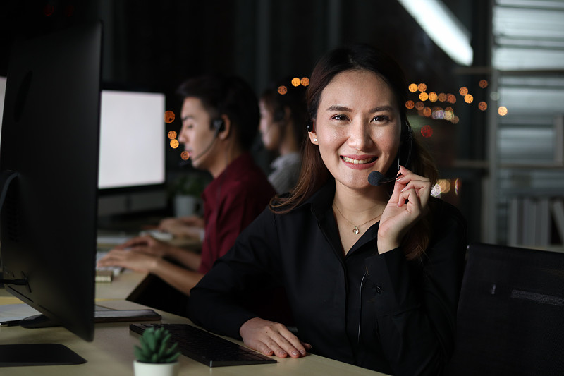 快乐微笑美女工作在呼叫中心服务台顾问与队友晚上，客户服务行政人员与麦克风耳机使用电脑支持在深夜的办公室。
