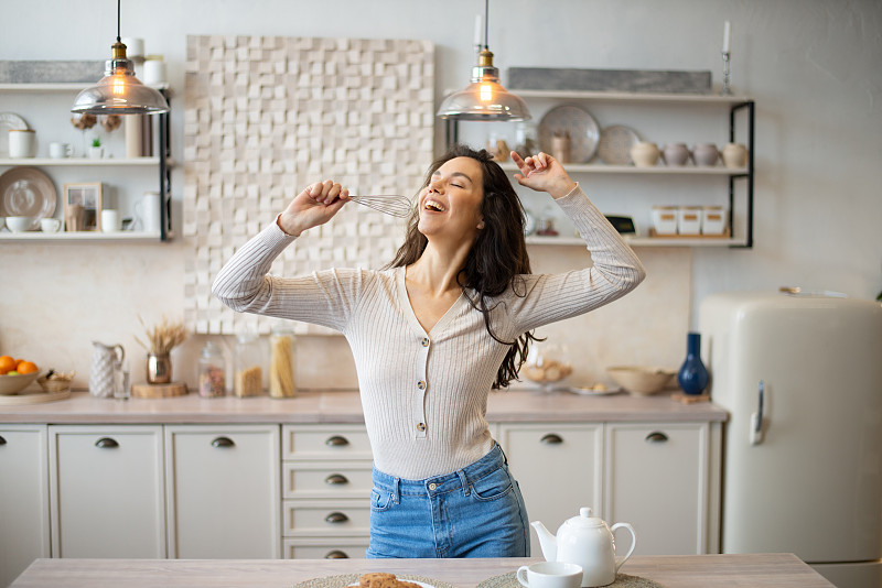 俏皮的年轻女子在器具上唱歌，就像想象中的麦克风，在明亮的厨房里跳舞，复制空间
