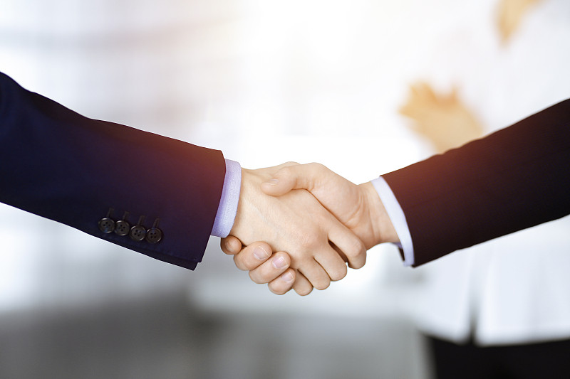 商务人士在会议或谈判中握手，特写。一群不知名的商人和一个女人站在一个阳光明媚的现代办公室里。团队合作，合作和握手的理念