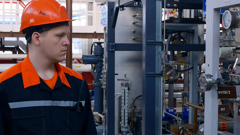 一名身穿橙色工作服、头戴安全帽的年轻工人在泵车间的化工厂的设备周围走动，检查设备的状况。工厂工作。