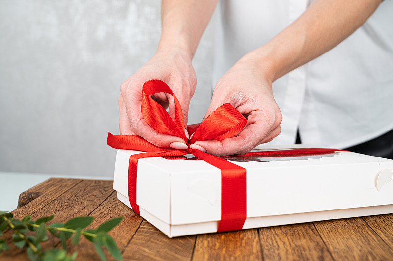 一个女人在一个白色的盒子上系一个红色缎子蝴蝶结的特写。礼物包装的概念