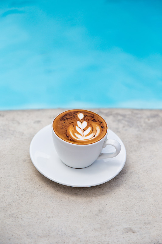 白色杯美味的卡布奇诺与拉花艺术靠近蓝色游泳池。