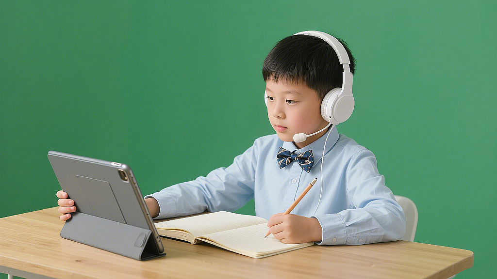 男孩正处于学习场景当中坐在一张木质的桌子前左手握持平板电脑